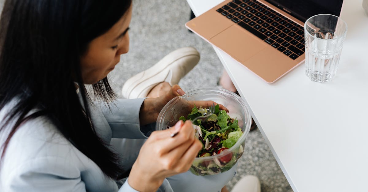 Woman eating a fresh salad at her desk with a laptop nearby, promoting healthy eating at work.