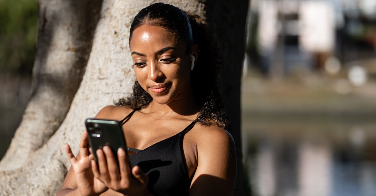 Smiling woman in activewear leans against tree while using smartphone outdoors.