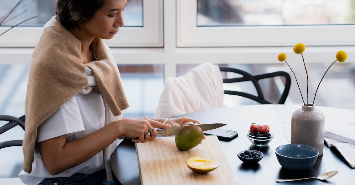 Woman slicing a mango indoors by a window, surrounded by bowls of fruit and kitchen items.