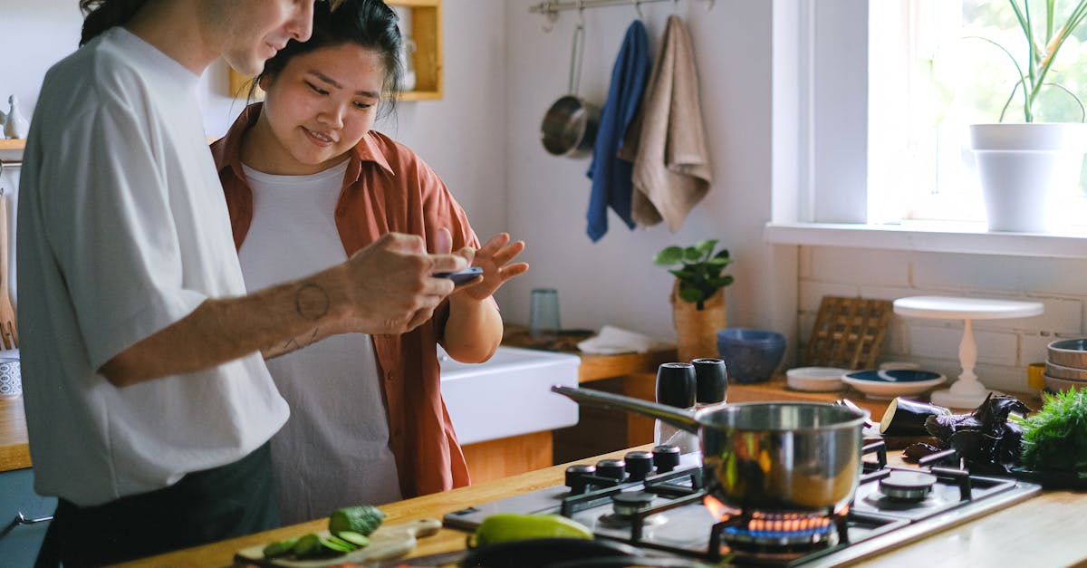 A young couple uses a phone while preparing a meal in their modern kitchen with fresh vegetables.