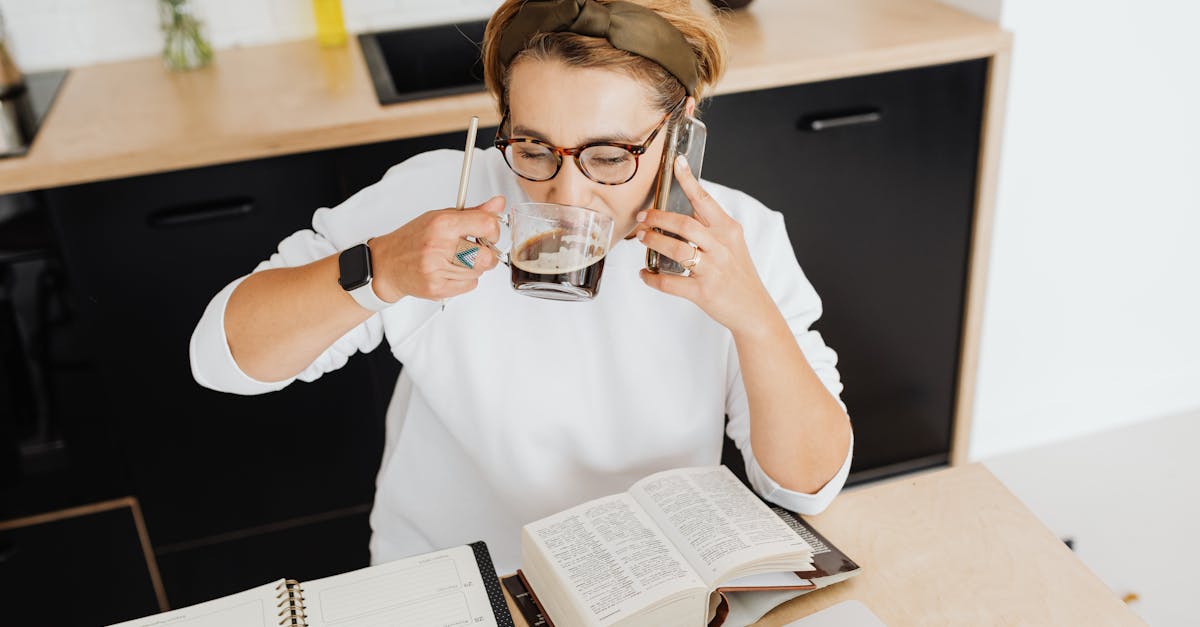 Adult woman drinking coffee, using smartphone, and studying at a kitchen table.