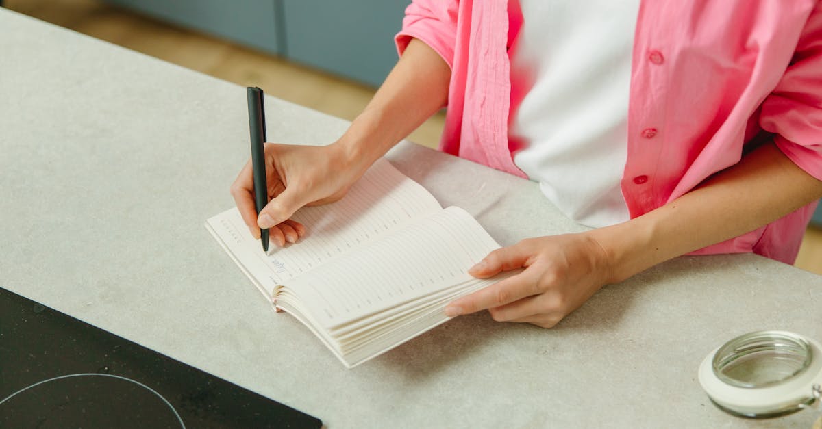 A woman in a pink shirt writing in a notebook on a kitchen counter with a pen.