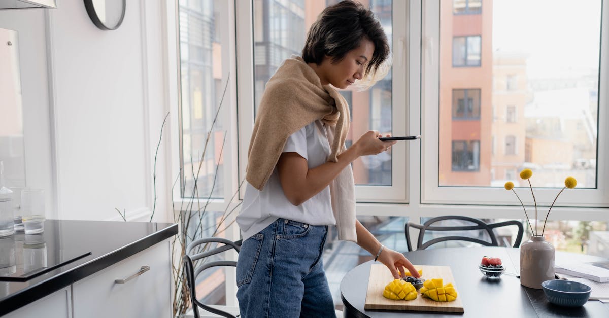 Young woman uses smartphone to photograph a cutting board with fresh mango in modern kitchen.